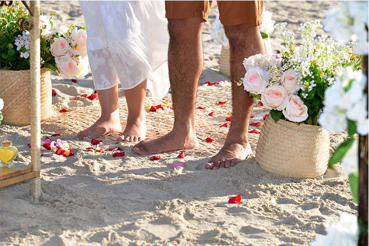 Pés de homem e mulher em cima de tapete de palha com pétalas de rosas na areia da praia