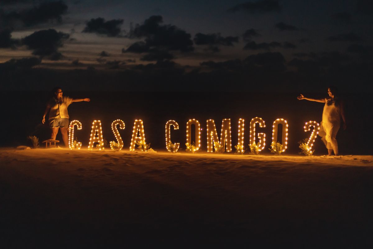 Duas mulheres com letreiro iluminado Casa Comigo? na praia no entardecer